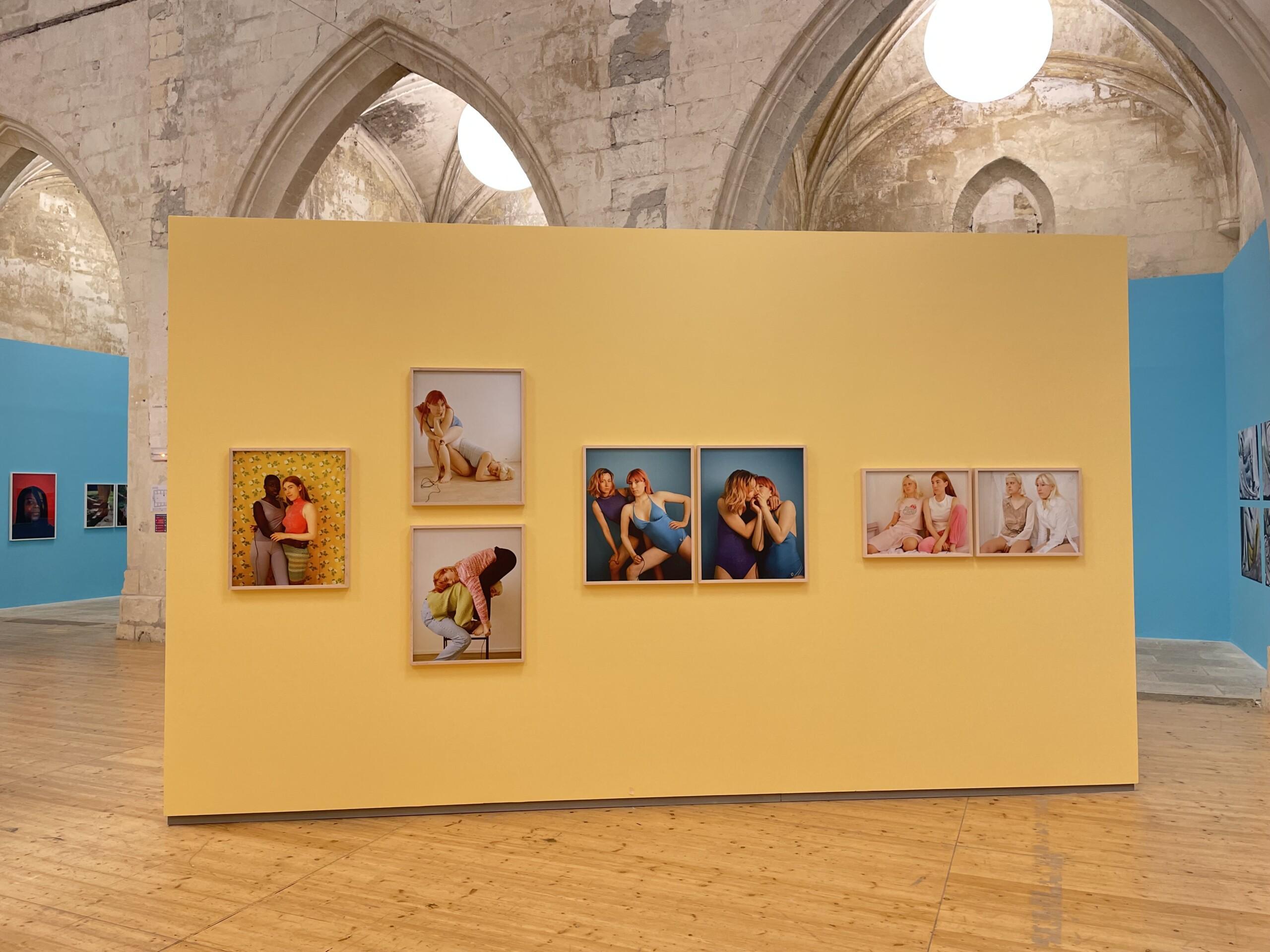 A yellow installation wall in the middle of a brightly lighted exhibition space. There are seven framed multi-colored photograph works by Emma Sarpaniemi installed on the wall.